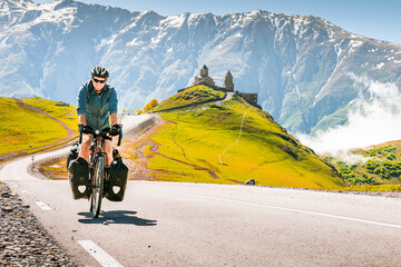 Cyclist back view on the road in scenic caucasus nature with Gergeti trinity monastery in the...
