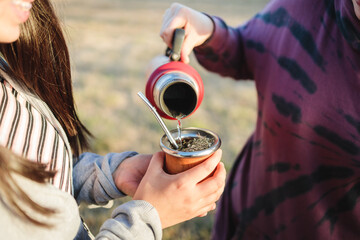 Two female friends brewing and drinking yerba mate, with a red thermos, in the countryside at sunset