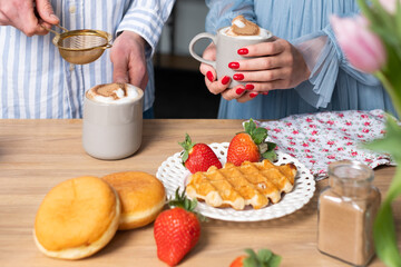 Obraz na płótnie Canvas A mans and a woman's hands in light blue clothes with red nails. A man ads cinnamon in a gray cup with caffe latte. The crispy waffle, red strawberries on a white plate and donuts.