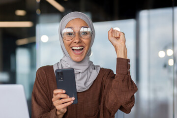 Closeup portrait of businesswoman in hijab, muslim celebrating victory and success good achievement results, workplace inside office looking at camera and happy, holding phone, received notification. - obrazy, fototapety, plakaty