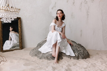 Elegant woman in dress sitting on big stone in studio full of sand