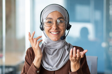 Online customer support worker consults a client remotely, a woman in a hijab with a headset for a video call talks, looks at the web camera and smiles at the interlocutor, closeup - obrazy, fototapety, plakaty