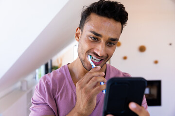 Happy biracial man brushing teeth and using smartphone in bathroom