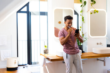 Happy biracial man brushing teeth and using smartphone in bathroom