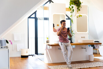 Happy biracial man brushing teeth and using smartphone in bathroom