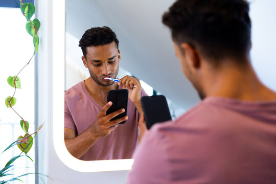 Biracial man in mirror, brushing teeth and using smartphone in bathroom - Powered by Adobe