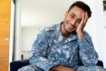 Happy biracial male soldier wearing uniform, sitting in armchair making video call at home