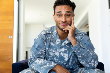 Happy biracial male soldier wearing uniform, sitting in armchair making video call at home