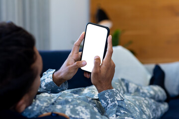 Biracial male soldier wearing uniform, lying on sofa using smartphone, with copy space on screen