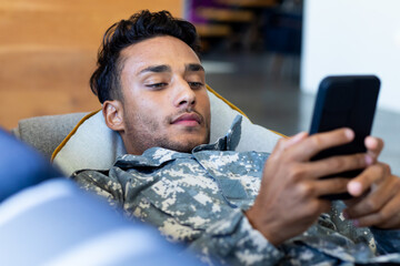 Biracial male soldier wearing uniform, lying on sofa using smartphone