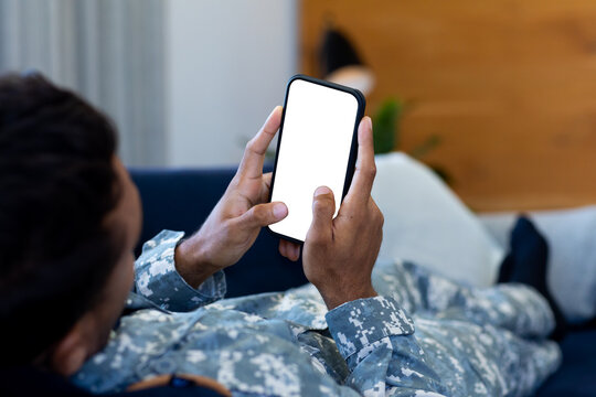 Biracial male soldier wearing uniform, lying on sofa using smartphone, with copy space on screen