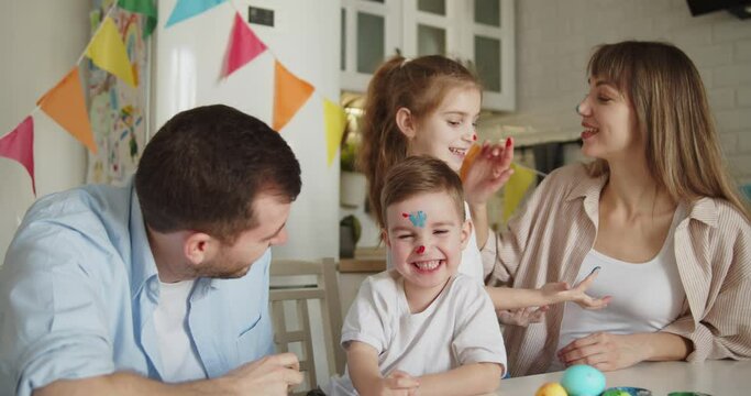 Joyful family of four painting Easter eggs together in bright modern kitchen. Parents and children fooling around drawing on face. Family gathering in preparation for Easter.