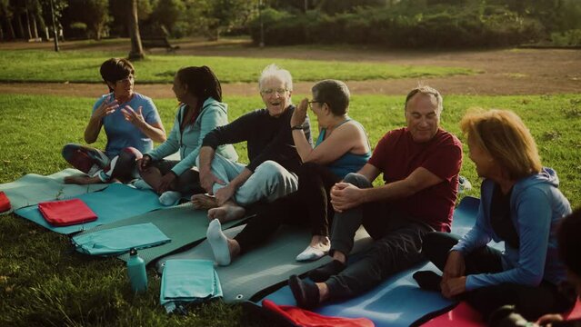 Group of multiracial senior friends taking a break sitting on mat after workout activity - Health elderly people lifestyle