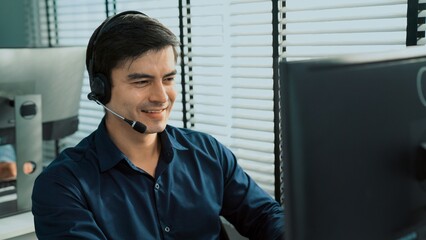 Young competent asian male call center agent working at his computer while simultaneously speaking...