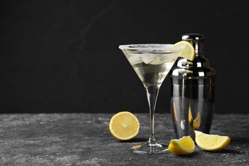 Martini glass of refreshing cocktail with lemon and ice cubes near shaker on black textured table. Space for text