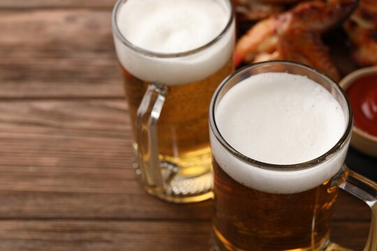 Mugs with beer and delicious baked chicken wings on wooden table, closeup. Space for text