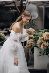 Side view of beautiful young man wearing wedding dress, posing outdoors. Attractive bride standing, smiling, looking at flowers, smiling. Concept of beauty and happiness.