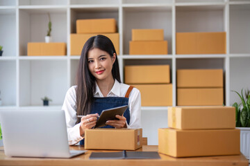 Obraz na płótnie Canvas A portrait of a young Asian woman, e-commerce employee sitting in the office full of packages in the background write note of orders and a calculator, for SME business ecommerce and delivery business