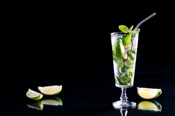 A tall Mojito cocktail with lime wedges on a glass table, against black.