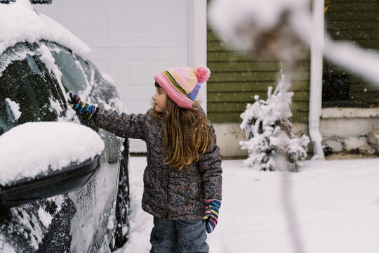 Happy Toddler Girl In Winter Gear Brushing Snow Off Car In Driveway