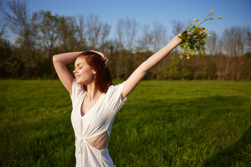 Naklejka na ściany i meble a beautiful, happy woman in a light dress stands in a field raising her hands high holding a bouquet of flowers