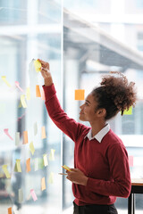 African American female employee write down on colorful sticky notes manage list, concentrated biracial woman work on startup brainstorm collaborate plan on stickers on glass wall