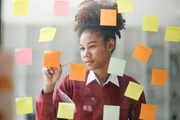 African American female employee write down on colorful sticky notes manage list, concentrated biracial woman work on startup brainstorm collaborate plan on stickers on glass wall