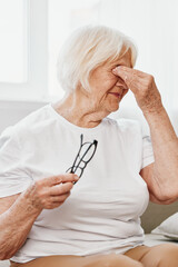 Elderly woman severe eye pain sitting on the couch, health problems in old age, poor quality of life. A grandmother with gray hair holds on to her sore eyes with glasses in her hand.