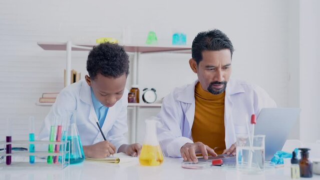 4K, African male student sits on a science experiment with a teacher teaching and guiding in his elementary school lab. Asian male is using laptop computer to check validity of  experimental results.
