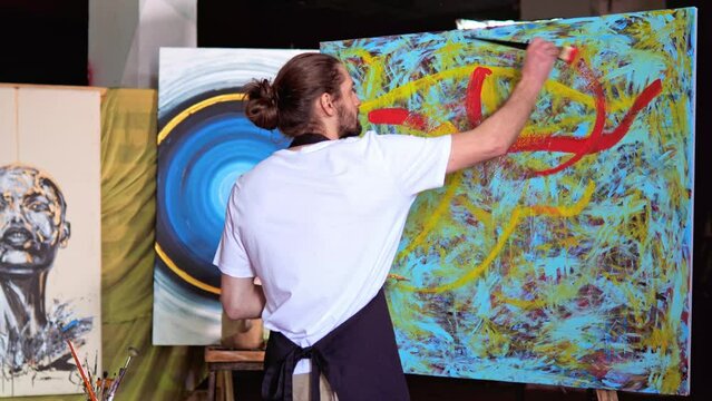 Portrait of talented artist working on abstract painting, using a brush to create emotional contemporary art on large canvas in creative studio. Man working up close and personal with their creation.