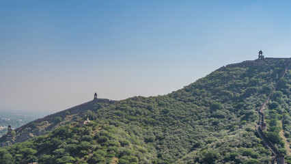 An ancient fortress wall runs along the top of the ridge of the mountain, overgrown with green trees. Watchtowers against the blue sky. India. Jaipur. Amber Fort