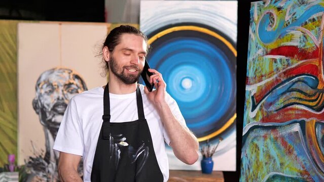 Handsome male artist stands in his studio holding cellphone in hand, in front of his abstract paintings. Man wearing a white t-shirt and is talking on his mobile phone while admiring his work.
