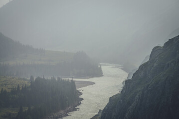 Misty mountain landscape with wide mountain river. Dark green gloomy scenery with confluence of two...