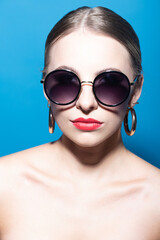 Fashion and make-up concept. Close-up beautiful woman portrait with classic hairstyle, big retro round sunglasses, red lipstick and golden round earrings in blue studio background