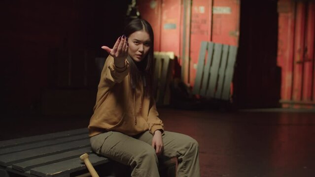 Aggressive tough pretty Asian female hooligan in street style clothes sitting on pallets, pointing with index finger and ordering to come near, expressing rudeness and hostility in abandoned building