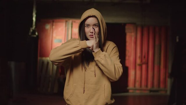 Aggressive rough beautiful Asian woman in hoodie showing intimidating fist gesture, threatening someone and expressing danger, bad attitude and anger in abandoned industrial premise.
