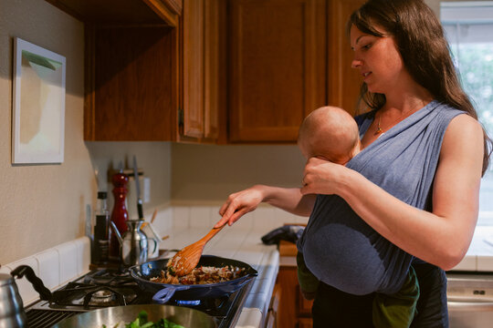 Mom holding newborn baby while cooking on skillet pan by range