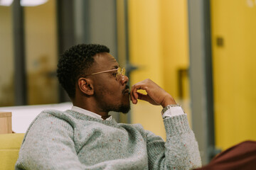 A side view photo of black male person sitting on a sofa at the office and thinking something