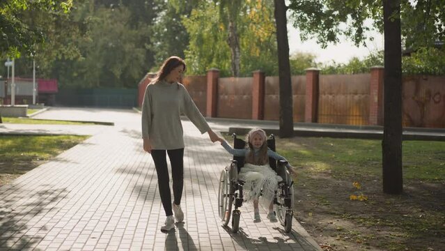 Woman walks on stone road holding hand of little daughter with muscular dystrophy. Caring mother spends time in park with little girl in wheelchair