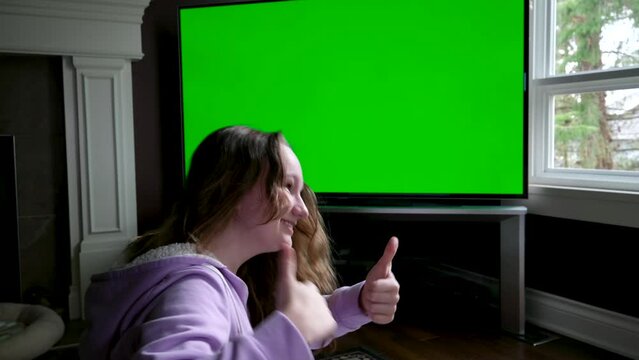 teenage girl sits admiringly in front of a big green chroma key tv monitor advertising space she enthusiastically points finger laughs thumbs up admiration open mouth wide say wow sincere emotions