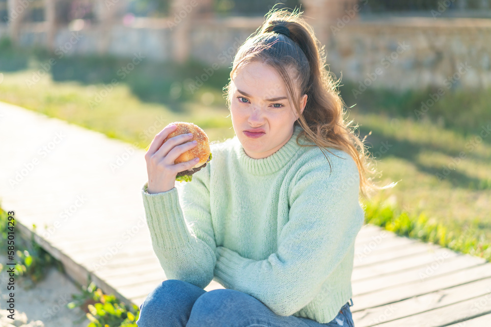 Poster Young pretty girl holding a burger at outdoors with sad expression