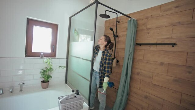 A Young Caucasian Woman In Rubber Gloves Cleaning A Shower Glass With A Sponge And Window Squeegee.