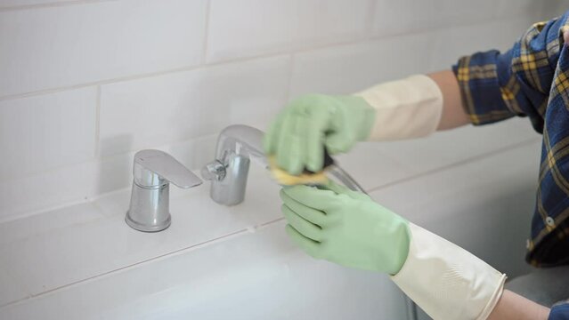 Close up shot of a person in rubber gloves cleaning a faucet using a sponge with bath detergent.