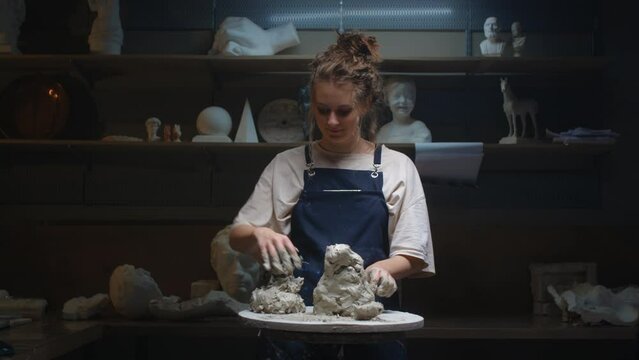 A young blonde European girl makes a sculpture of white clay with her hands, creates a work of art statue for the interior. A woman touches the soft clay on a potter's wheel with her fingers.