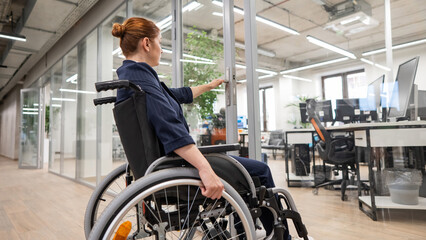 Red-haired caucasian woman in a wheelchair trying to open the door in the office. 