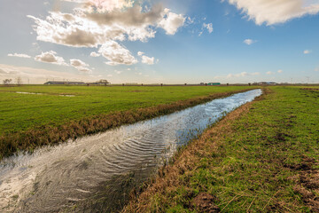 Fototapeta na wymiar Turbulent water surface in a ditch. The photo was taken at the end of the winter season on a stormy day in the Dutch province of North Brabant. In the background are a barn and a row of wind turbines.