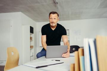 Mature Businessman Sitting on Desk in Office with Laptop and Looking Thoughtful at Something Off-Camera