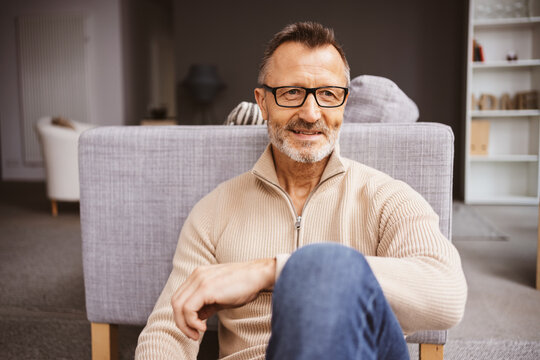 Relaxed 50-Year-Old Man with Glasses Sitting Comfortably on the Floor in Front of His Sofa and Smiling at Something Off-Camera