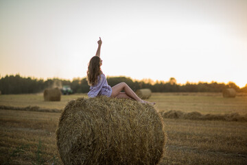 Naklejka na ściany i meble a happy girl with long hair sits on a haystack in a field at sunset and enjoys nature, loneliness and freedom.