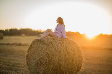 Naklejka na ściany i meble a happy girl with long hair sits on a haystack in a field at sunset and enjoys nature, loneliness and freedom.